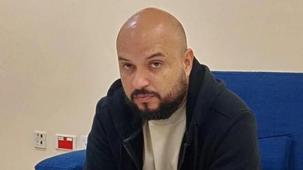 A head and shoulders image of Arturo Carrera Loaiza wearing a dark, warm jacket and white top. He is sitting on a blue chair.