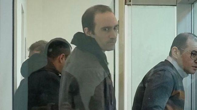 A screengrab from a video shows a man in a hoodie standing with short hair and a jacket and looking at the camera