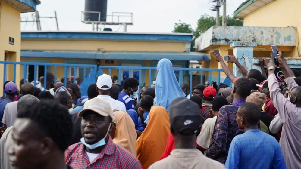 A crowd of people gather outside Minna hospital. You can see the backs of their heads and many are wearing caps. They stand in front of a blue metal gate, behind which sits yellow buildings with blue detailing.