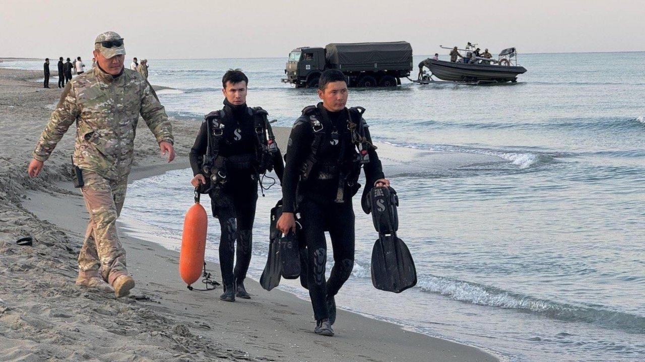 Three men walk on a coast - one is wearing army camouflage uniform, the other two what appear to be black diving kits carrying flippers and an orange buoy. Behind them, a military lorry appears to be towing a boat with military personnel on board.