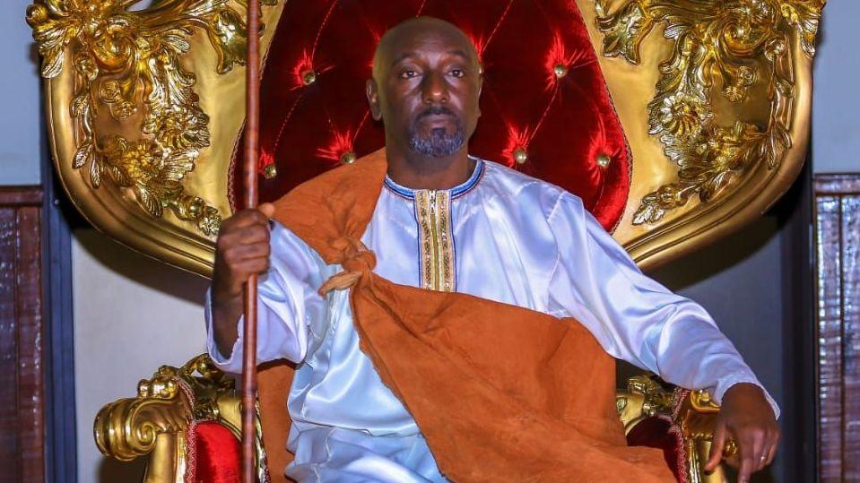 Edward Rukidi Kijanangoma wearing traditional clothing of a white gown with an orange cloth tied around the neck and holding a spear