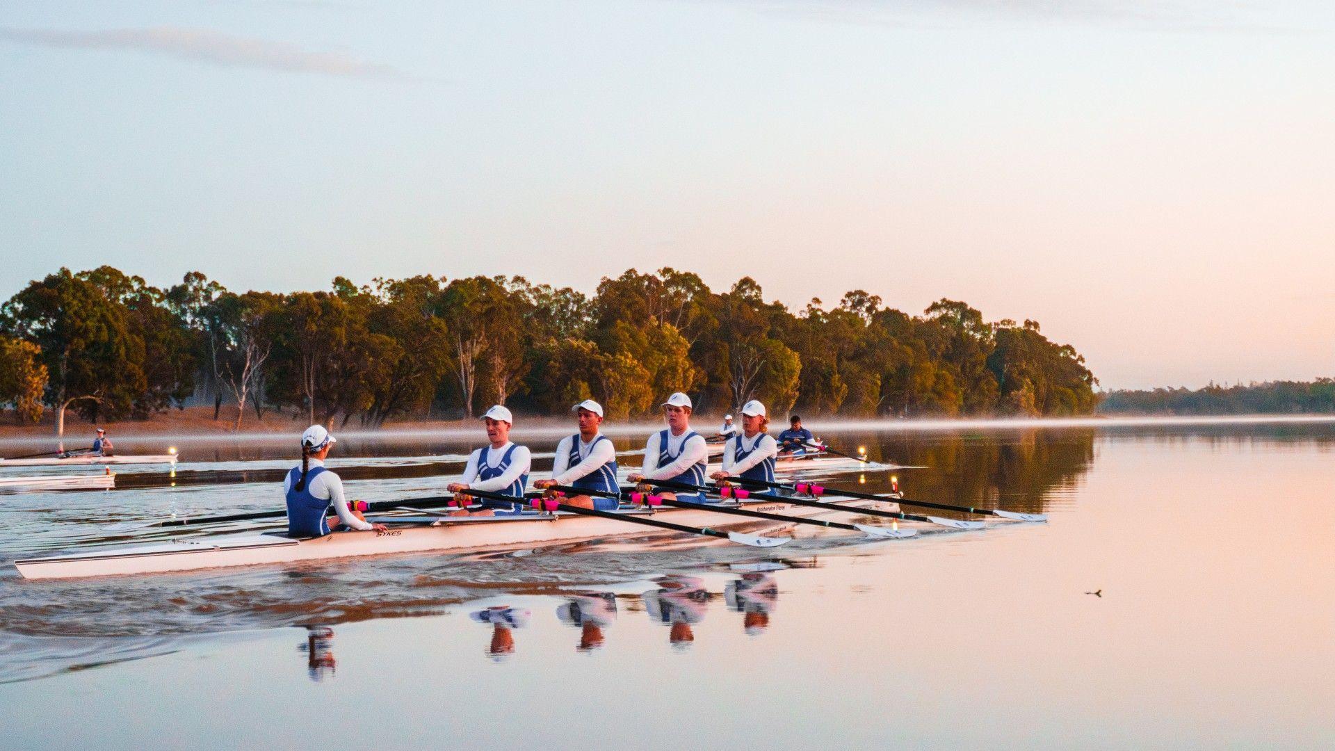 A rowing boat with rowers wearing white and blue row down a wide river with mist rising from the water.