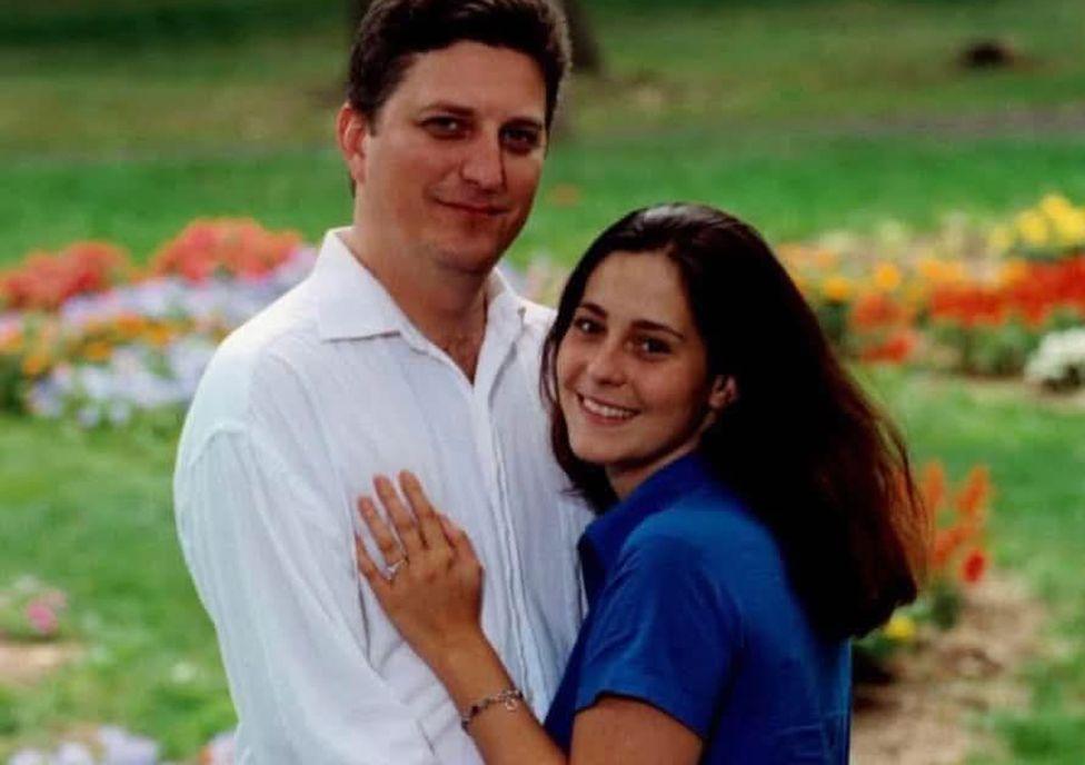 John Resta is on the left, he has dark hair and is wearing a white linen shirt and his wife Sylvia is on the right, with dark long hair and a blue short-sleeved shirt. She is pressed against him, her left hand on his chest. It has a wedding ring on it. They are both smiling. Behind them is grass with flowers.