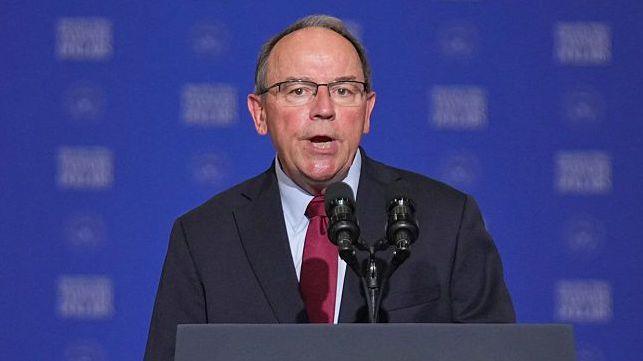 Close-up of Tom Tiffany, wearing a suit and tie, speaking into a microphone at a podium in front of a blue background