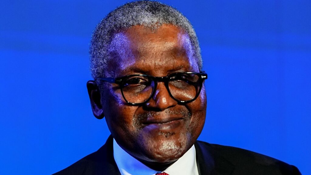 A man with grey hair, wearing a black suit, white shirt, and red tie, smiling into the camera.