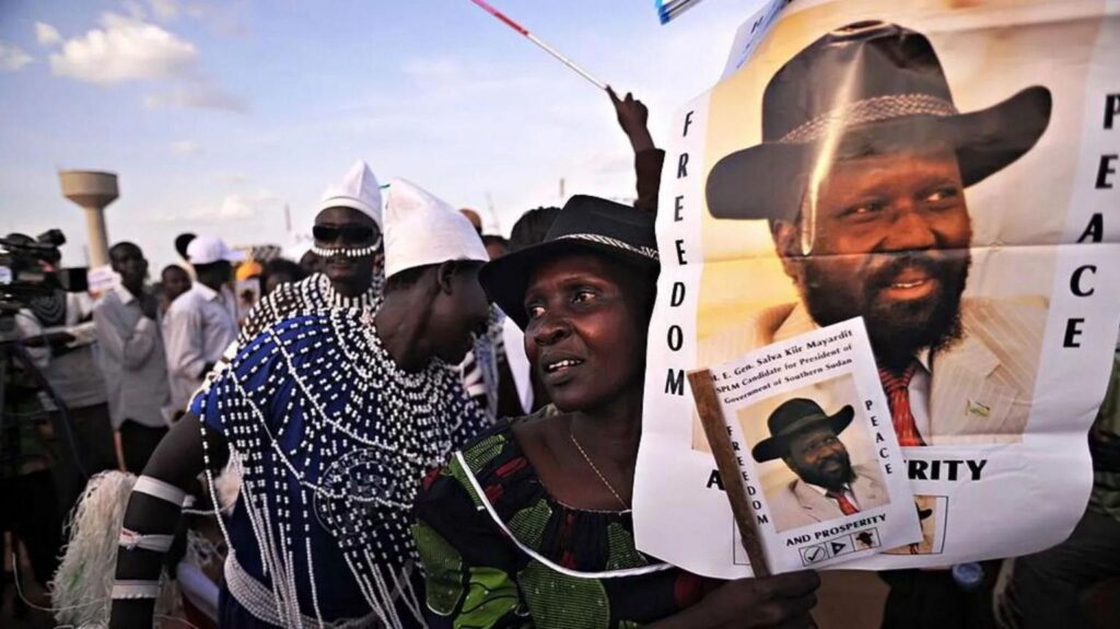 A crowd at a political rally holding a banner for South Sudan President Salva Kiir that reads "Freedom, peace and prosperity next to his portrait wearing a black cowboy hat