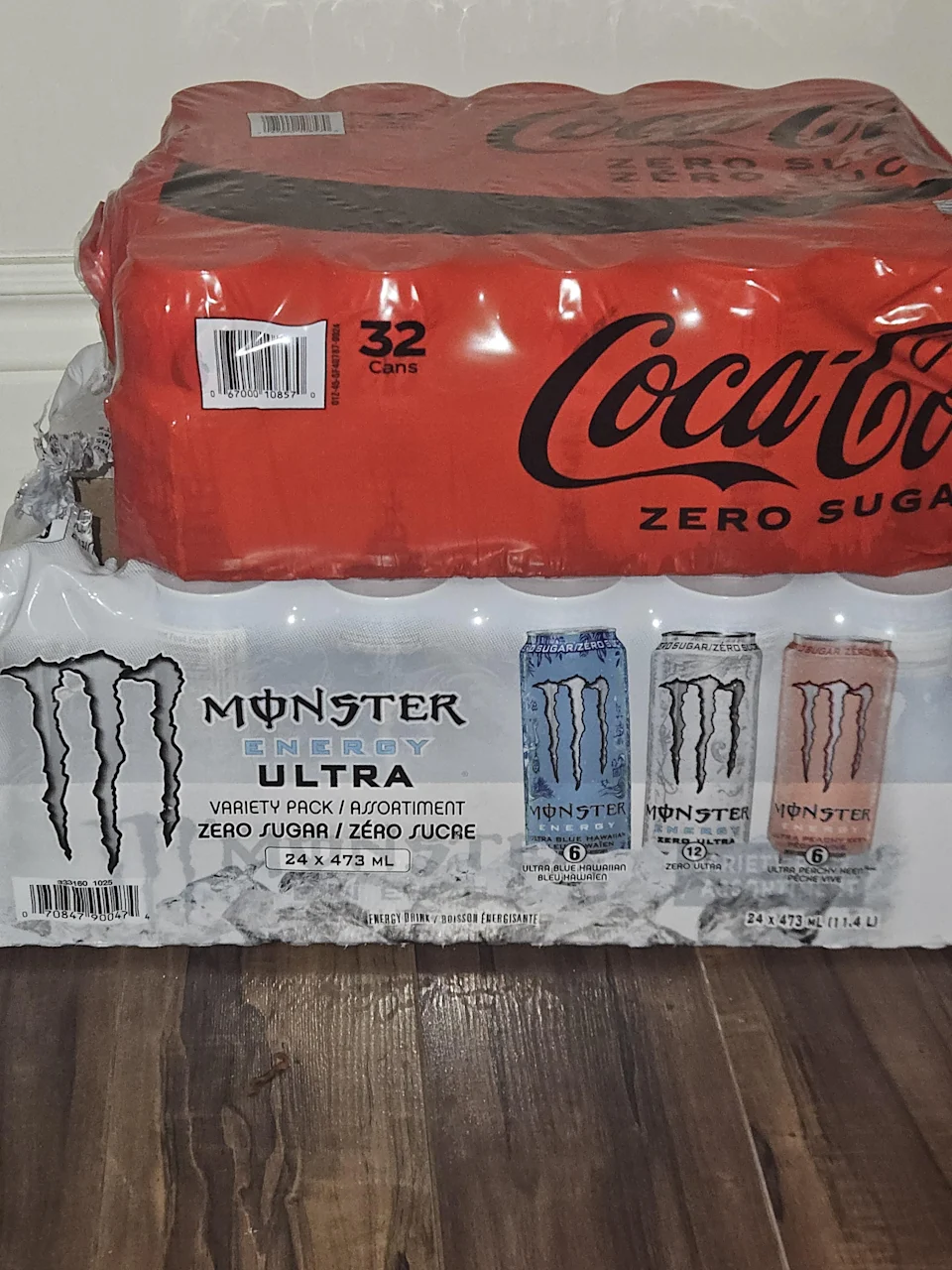 Cases of Coca-Cola Zero Sugar and Monster Energy Ultra in a variety pack are stacked on a wooden floor