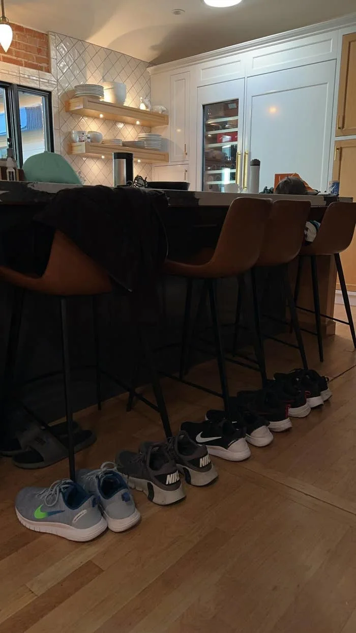 Five pairs of sneakers are lined up on the floor in front of a kitchen island with barstools and a modern kitchen setup in the background