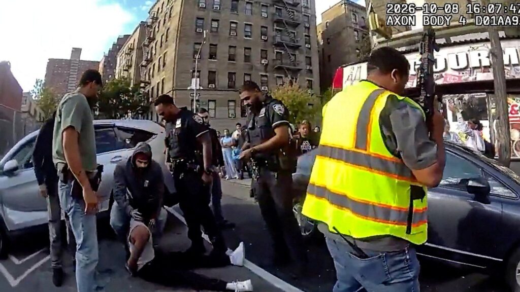Footage from a New York police officer's body camera shows a man sat on the ground by a silver car with four other men, including police officers, stood around him. One man, wearing a hooded jumper, bends over him and touches his neck. A man holding a large gun and wearing a high-visibility vest is seen in the foreground on the right of the shot, in front of a dark car