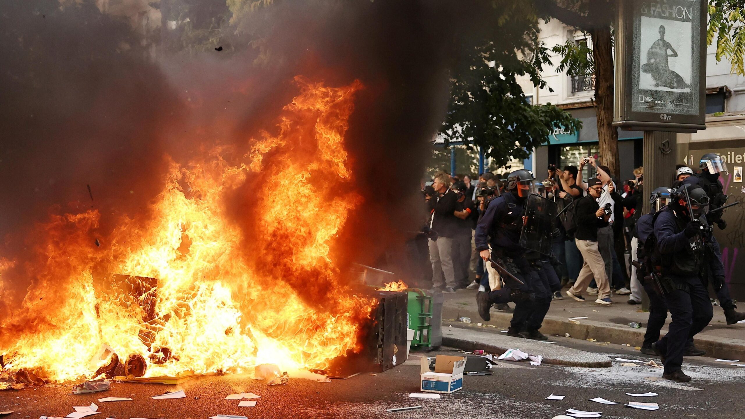 A large fire burns on a road in Paris beside which five officers in riot gear walk past a crowd observing the flames. There is debris strewn across the street.