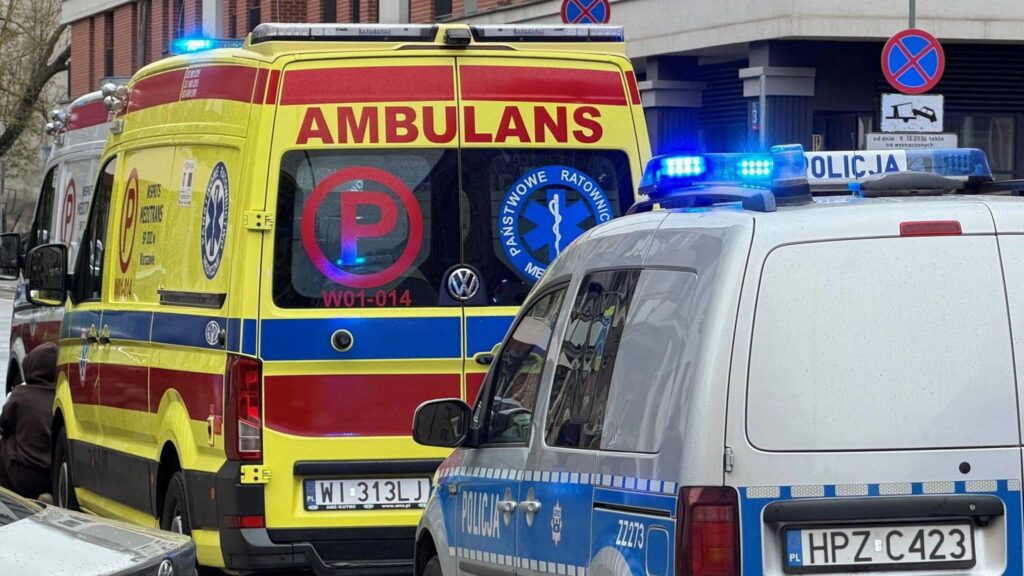 An ambulance of the Polish ambulance service parked in front of a grey Polish National Police van on the side of the road, both have blue sirens on