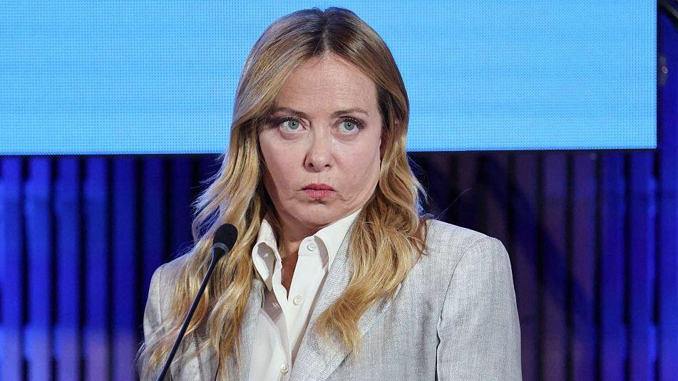 Giorgia Meloni pictured speaking on stage. She has long blonde hair and wears a grey blazer and white buttoned shirt. She stands in front of a small black microphone, with a bright blue screen behind her.
