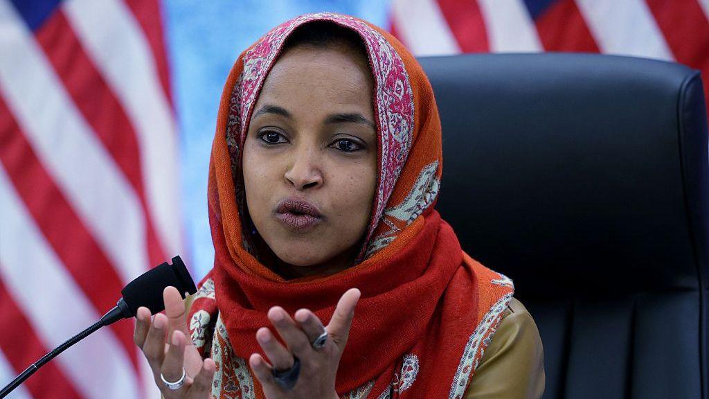 Ilhan Omar gestures as she speaks. She is seated in a black chair and wearing an orange-coloured patterned headcovering.