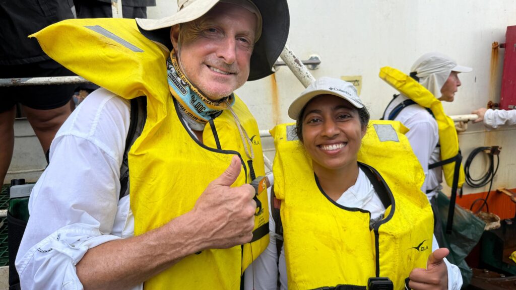 Expedition members Steve Schultz and Sirisha Bandla give a thumbs up and smile to the camera while wearing yellow life vests and white tops on the deck of a ship. Schultz wears a cream bucket hat and Bandla a cap.