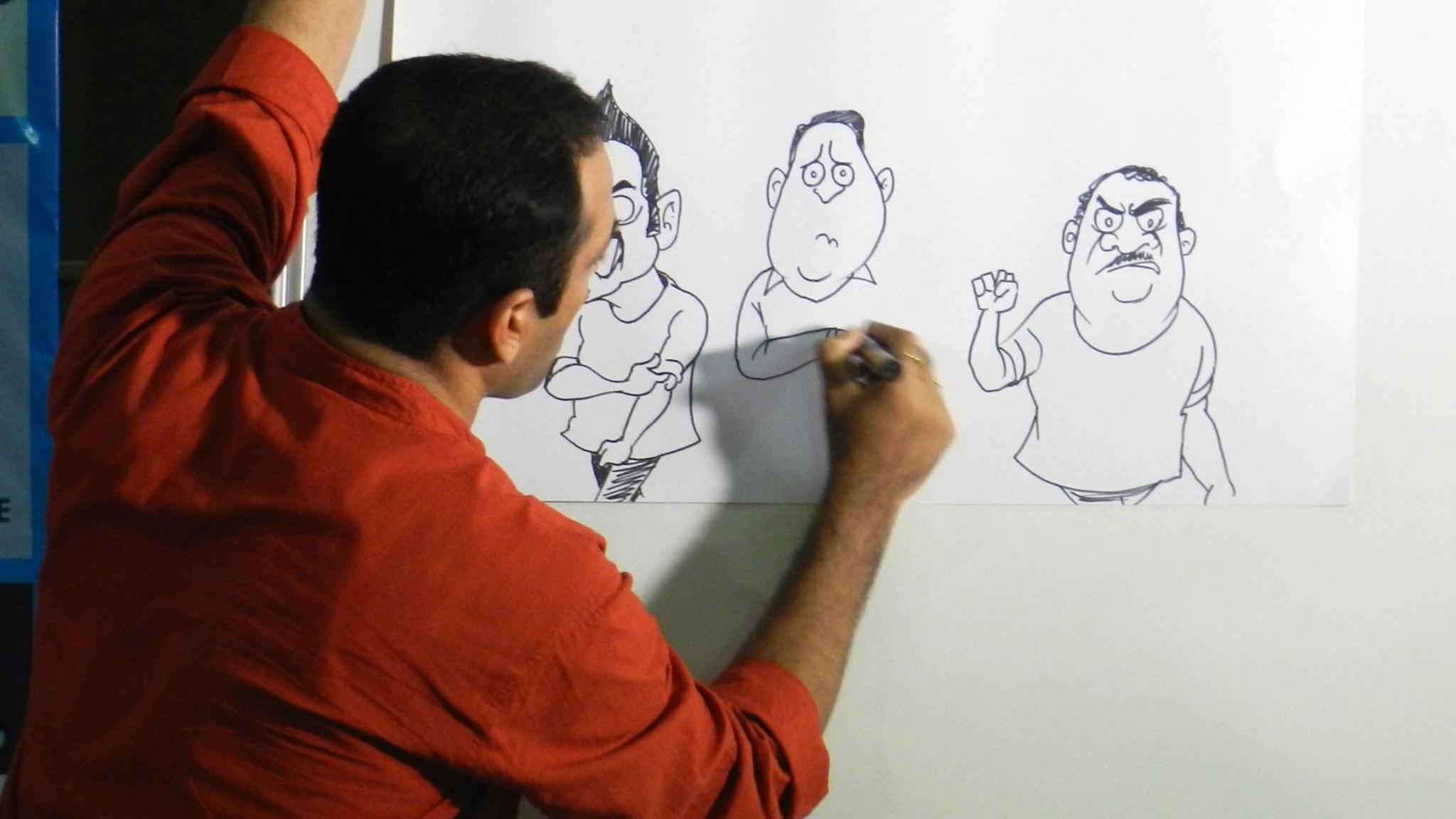 Satish Acharya wearing a rust shirt draws cartoons on a piece of paper attached to a white board
