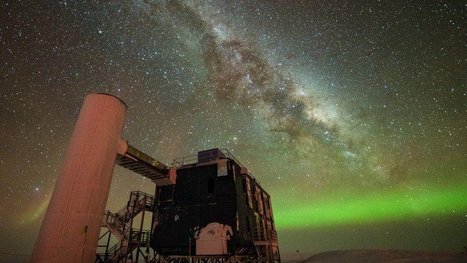A starry sky the Milky Way is seen through the middle, and the Northern Lights in green along the horizon. In the foreground is an observatory with a black building and stairs liking to a white column.
