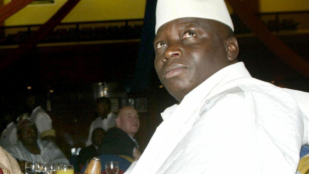 Former President Yahya Jammeh of Gambia seated in a white robe seated at a table.
