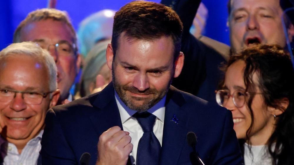 Paul St-Pierre Plamondon smiles and clenches his fist in victory. He is surrounded in the background by smiling supporters.