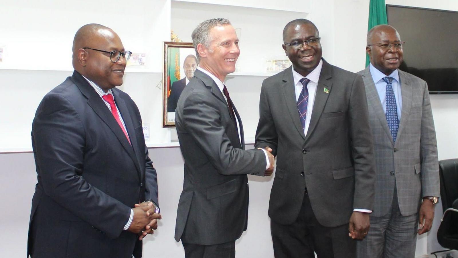 Four men stand in suits, two shaking hands and smiling in the middle.