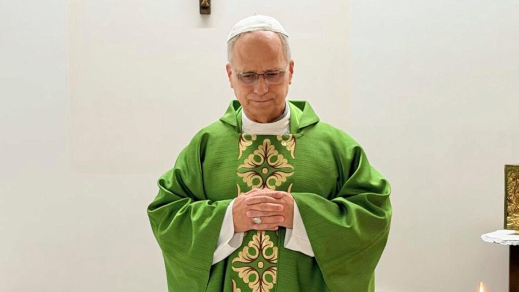 Pope Leo celebrates mass from hospital, wearing a green robe and standing before a white wall adorned with a wooden crucifix.