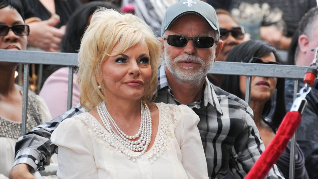 Televangelist Jim Bakker and wife Lori Bakker sitting at a Hollywood Walk of Fame ceremony in October 2011. Jim Bakker is looking forward at the camera with sunglasses, wearing a cap with a cross and a plaid shirt. Lori Bakker is looking forward and smiling, with a white blouse and pearls