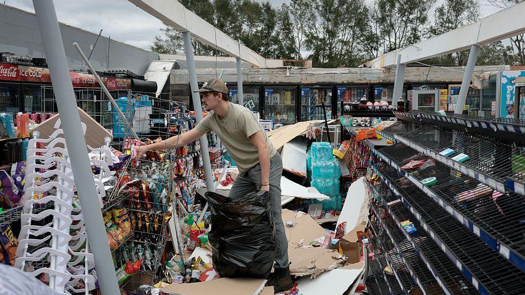 A man puts rubbish in a bag with debris strewn all around a store with the roof blown off