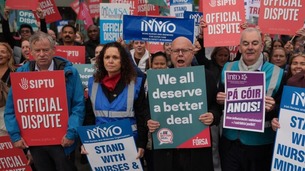 Three men and a woman stand at the forefront of a large protesting crowd. The four people in front are wearing luminous coats and are holding placards. The crowd behind are also holding a range of placards.