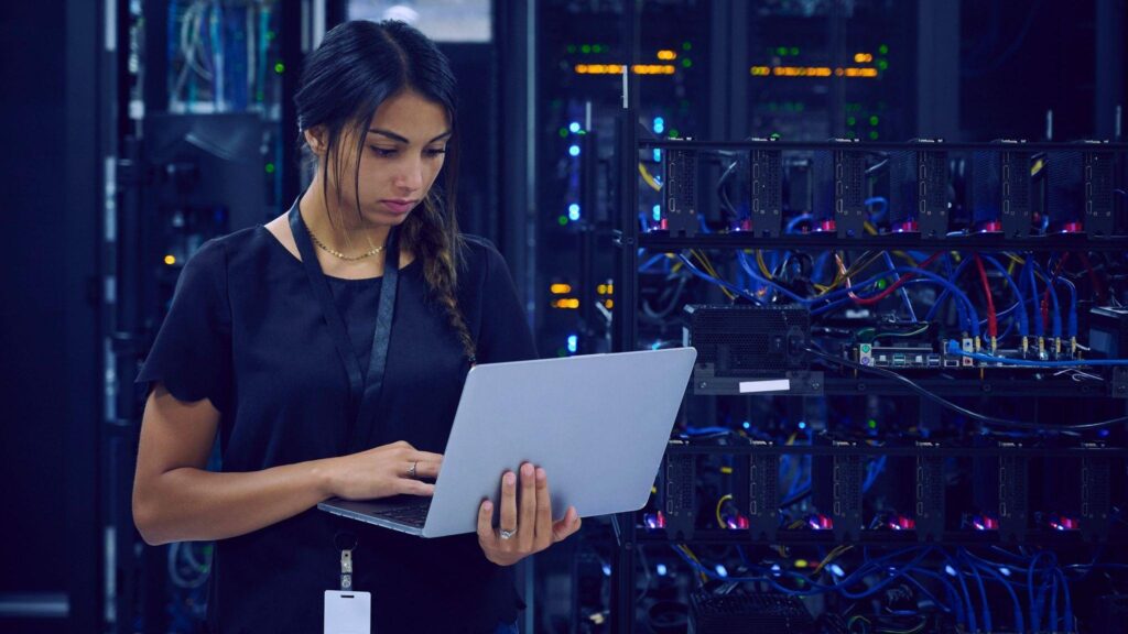 A female technician dressed in navy blue clothes and wearing a lanyard around her neck is standing in a server room using her laptop.