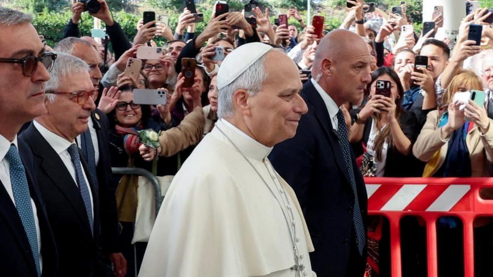 The Pope as he arrives at the hospital, with security following him and dozens of people behind barriers holding their phone up to catch a photo of him
