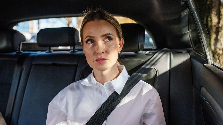 A woman wearing a white shirt sits in the back of a car. She is looking out the window and is waring a seatbelt. She doesn't look happy.