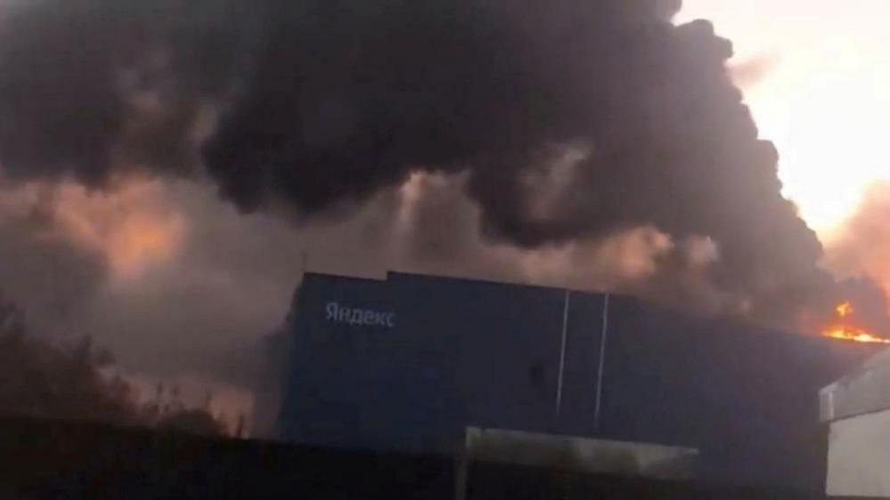 Large warehouse building engulfed in flames as thick black smoke billows into the sky