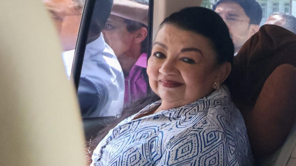 Shiranthi Rajapaksa, seated inside a vehicle, looks toward the camera. She is wearing a white shirt with a blue geometric pattern, while other people are visible on the street through the car windows
