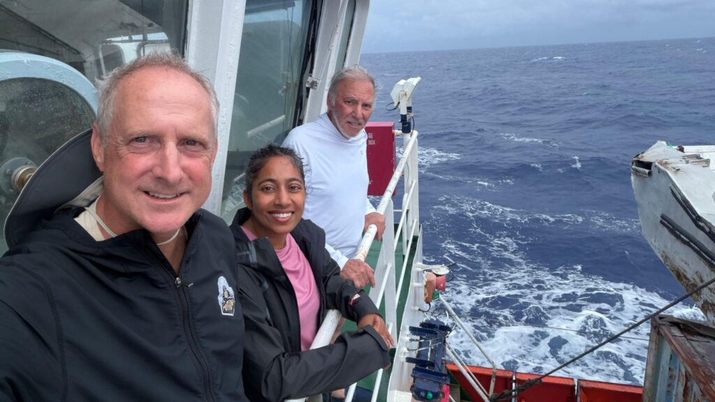 Two men and a woman stand on the viewing deck of a vessel travelling through the Pacific Ocean.
