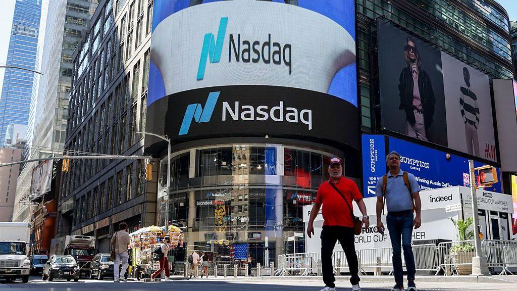 Two men walk in front of the Nasdaq building, which has a large curved lit up sign that reads Nasdaq on the front.