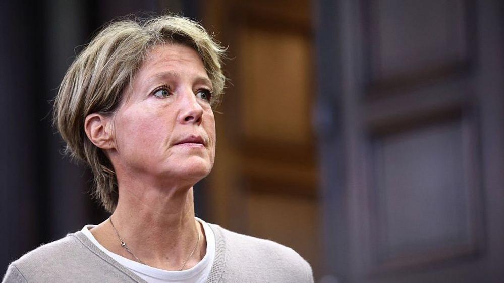 A woman with short blonde hair and a beige sweater and white T-shirt looks to the right