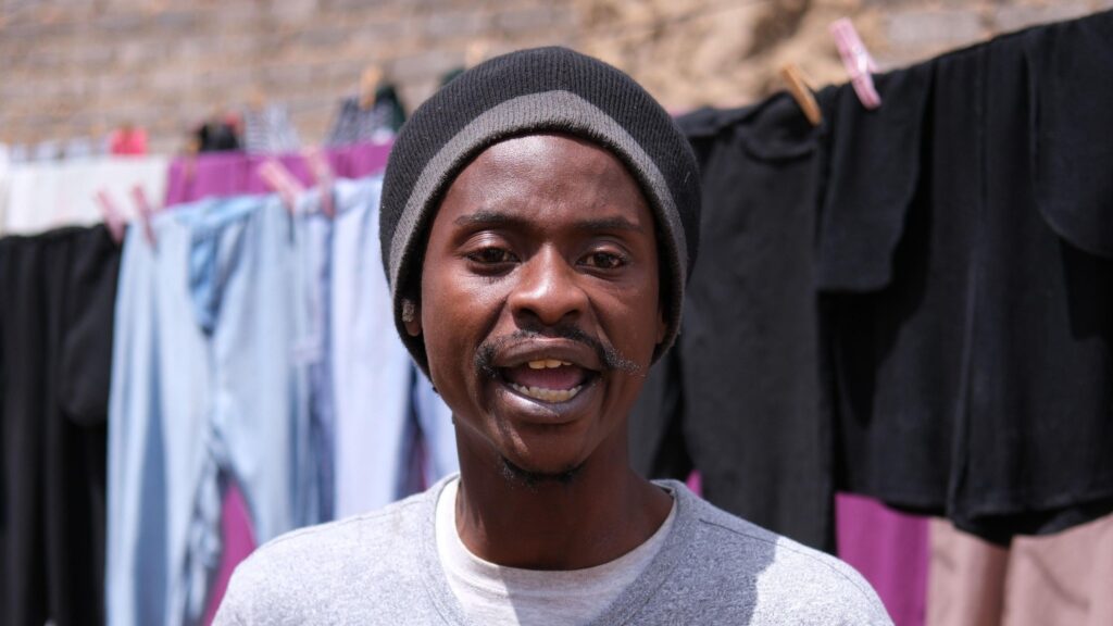 A close up short of a man speaking looking directly forward. He has a short beard, is wearing a dark grey long-sleeve shirt. The background features dark laundry hanging out to dry on a clothesline.